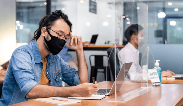 An employee works with a screen guard and a mask on in his office which has hygiene protocols implemented